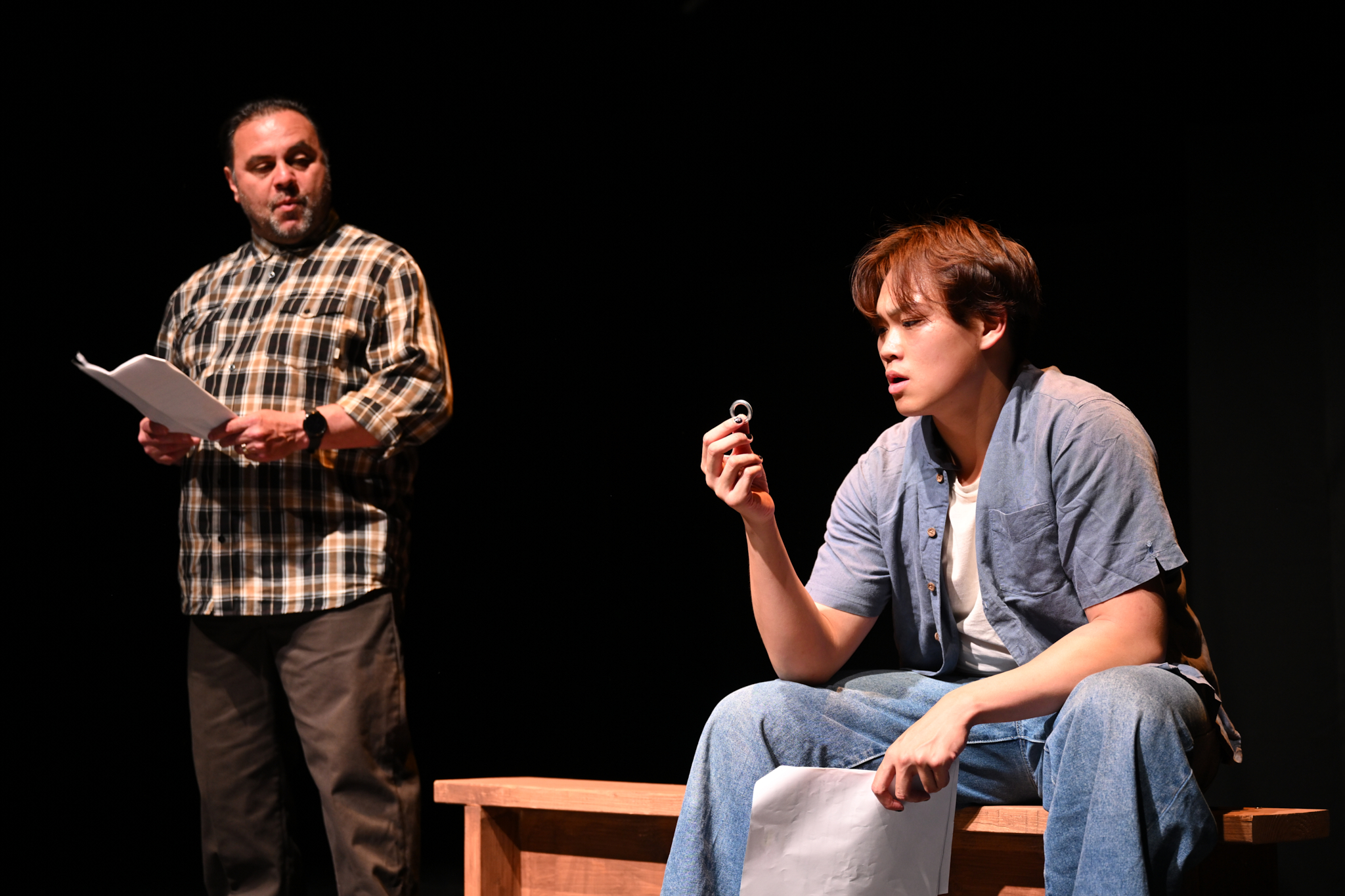 Photo from Beyond Prison Walls 2024 at SDSU. DESCRIPTION: Man standing up and holding script looks down at young man sitting down and looking at a ring in his hand.